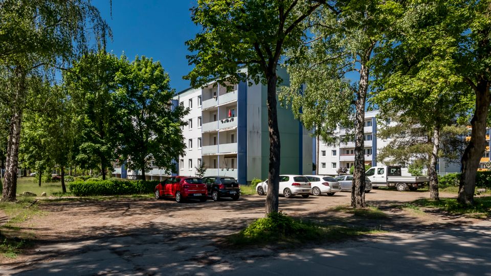 Wohnsiedlung mit mehrstöckigen, hellen Mehrfamilienhäusern und Balkonen, umgeben von hohen Laubbäumen. Im Vordergrund liegt ein kleiner Parkplatz mit mehreren Autos und eine asphaltierte Zufahrt zwischen Grünflächen.