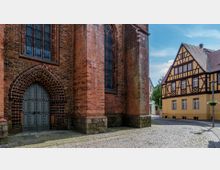 Eine gotische Kirche mit Backsteinmauerwerk und Spitzbogenportal steht neben einem Fachwerkhaus mit gelber Fassade und braunen Holzdetails. Der gepflasterte Platz im Vordergrund verbindet die beiden Gebäude, die von blauem Himmel und einigen Bäumen umrahmt sind.