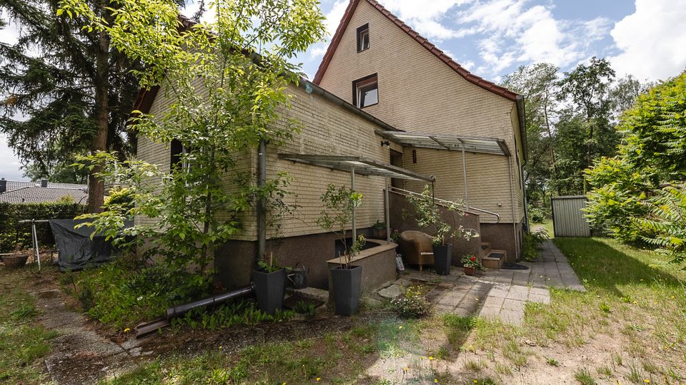 Seitenansicht eines zweigeschossigen Wohnhauses mit hell verklinkerter Fassade und steilem Satteldach, neben einem kleinen gepflasterten Terrassenbereich mit Überdachung und Stufen. Umgeben von Bäumen, Sträuchern und Rasen führt ein schmaler Weg zur Rückseite, daneben steht ein kleiner Schuppen.