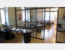 Ein offenes Büro mit Glaswänden, die Arbeitsbereiche abtrennen. Der Raum verfügt über Holzböden, Schreibtische mit Computern, Bürostühlen und Arbeitsmaterialien sowie eingeschränkte Dekoration an den Wänden.