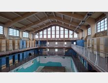 Innenansicht eines alten, leeren Schwimmbads mit bröckelnden Wänden, einem leeren Becken und einer umlaufenden Galerie mit Geländern. Große Fensterreihen lassen Tageslicht in den Raum, während Umkleidekabinen mit abgenutzten Türen die Wände säumen.