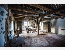 Historischer Innenraum eines Bauernhauses mit Sichtbalkendecke, gemauerten Wänden aus Ziegeln und Feldsteinen, sowie einem rustikalen Essbereich mit Holztisch und Stühlen. Der Raum ist mit Fliesenboden, einem Holzschrank und Fenstern mit Sprossen ausgestattet.