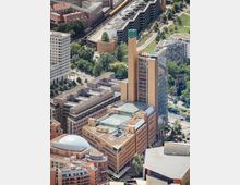 Aerial view of a large urban office complex with tan modern buildings, a tall narrow tower topped with green glass, and a taller dark glass high-rise beside it. Surrounding streets, trees, and adjacent buildings—including rail tracks and a green park area—frame the site.