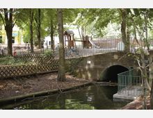 Spielplatz in einem Park mit hohen Bäumen, im Hintergrund ein hölzernes Klettergerüst mit Rutsche. Im Vordergrund eine kleine, mit Efeu bewachsene Brücke über einen Bach, daneben ein Zaun aus Holz und Metallgeländer.