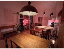 Gemütlicher Wohnraum mit Esstisch aus Holz, einer Küche im Hintergrund und einer großen Hängelampe als zentrale Beleuchtung. Im Raum befinden sich Pflanzen, ein Regal mit Weinflaschen, offene Holzstühle und rötliche Akzentbeleuchtung, die eine warme Atmosphäre schafft.