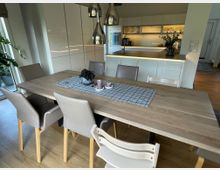 Essbereich mit hellem Holztisch, umgeben von beigefarbenen Stühlen und einer Kinderstuhlanordnung, angrenzend an eine moderne, offene Küche mit weißen Hochglanzschränken und einer beleuchteten Arbeitsplatte. Im Hintergrund sind eine große Fensterfront sowie Zimmerpflanzen sichtbar.