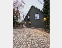 Ein modernes Einfamilienhaus mit dunkelgrauer Fassade und großen Fenstern, umgeben von gepflastertem Hof und Bäumen; im Hintergrund ist ein Zaun sichtbar.