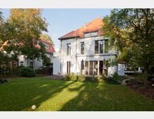 Zweistöckiges Einfamilienhaus mit weißer Fassade und rotem Satteldach, umgeben von einem gepflegten Garten mit Rasen und Bäumen. Große Fenster und eine Terrasse mit Sonnenschirmen prägen die Rückseite des Hauses.