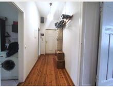 Narrow apartment hallway with white walls and a wooden plank floor leading to a white door at the end, lit by a hanging ceiling lamp. A wall-mounted rack with hangers and a few cardboard boxes lines the right side, and an open doorway on the left reveals a small laundry area with a washing machine.