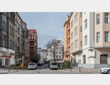Straßenszene in einem Wohngebiet mit mehrstöckigen Altbauten auf beiden Seiten, einige in hellen Farbtönen und andere in dunklerem Grau. Geparkte Autos säumen die ruhige Straße, und im Hintergrund sind weitere Gebäude und kahle Bäume erkennbar, die auf die Winter- oder Frühjahrszeit hindeuten.