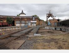 Historisches Eisenbahndepot mit einer Drehscheibe in der Mitte, umgeben von Backsteingebäuden mit Garagentoren und einem runden Turm. Im Hintergrund sind ein alter Wasserturm, Schienenfahrzeuge und technische Anlagen sichtbar.