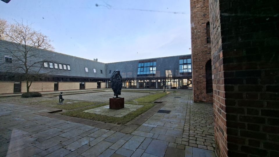 Innenhof eines Gebäudekomplexes mit gepflasterten Wegen, Rasenflächen und einer zentralen Skulptur auf einem Sockel. Umgeben von flachen Gebäudeteilen mit dunkler Fassadenverkleidung und Fensterbändern sowie einem hohen Backsteinbau an der rechten Seite.