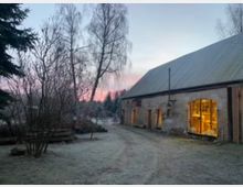 Ein frostiger Hof mit einer alten Scheune aus Ziegelstein, deren erleuchtete Fenster Einblick in eine Werkstatt gewähren. Im Hintergrund sind kahle Bäume vor einem Himmel mit rosafarbenem Sonnenaufgang zu sehen.