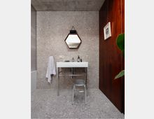 Modern bathroom vanity area with terrazzo walls and floor, featuring a white rectangular sink on chrome legs, a hexagonal mirror, and a towel hanging beside the basin. A dark wood-paneled wall with a framed picture contrasts with the concrete ceiling and a small metal stool in front of the sink.