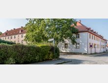 Straßenecke in einem Wohngebiet mit gepflastertem Gehweg und Straße, umgeben von bepflanzten Hecken. Im Hintergrund stehen Gebäude mit roten Ziegeldächern und heller Putzfassade, ein großes grünes Blattwerk eines Baumes ragt ins Bild. Ein Straßenschild zeigt „Friedrich-Ebert-Str.“.