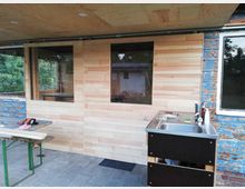 Holzverkleidete Außenwand mit großen Fenstern, daneben eine freistehende Spüle aus Metall. Im Vordergrund ein Klapptisch mit Werkzeugen, im Hintergrund eine Gartenlandschaft und ein Nebengebäude.