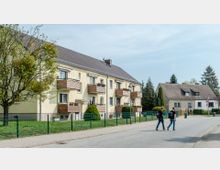 Mehrfamilienhaus mit gelber Fassade und mehreren Balkonen, umgeben von einem grünen Garten mit einem Metallzaun. Daneben stehen Einfamilienhäuser, und auf der Straße im Vordergrund gehen zwei Menschen.