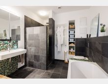 Modernes Badezimmer mit Dusche, Badewanne und Waschbecken. Die Wände und der Boden sind mit dunklen Fliesen verkleidet, ergänzt durch grüne Mosaikakzente; ein Handtuchheizkörper und Regale mit Körben befinden sich an der Wand.