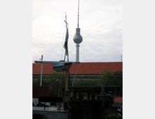 Ein Blick auf den Berliner Fernsehturm vor bewölktem Himmel, im Vordergrund ein Boot mit Masten und einer roten Ziegeldach-Kulisse im Hintergrund.