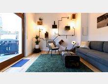 Modern eingerichtetes Wohnzimmer mit einer grauen Couch, einem Sessel und einem Couchtisch aus Metall in Kofferoptik. An der weißen Wand befindet sich eine sichtbare Rohrinstallation mit Pflanzen, Lampen und Dekorationsgegenständen, daneben steht eine große Glastür mit Blick auf einen Außenbereich mit Grill.