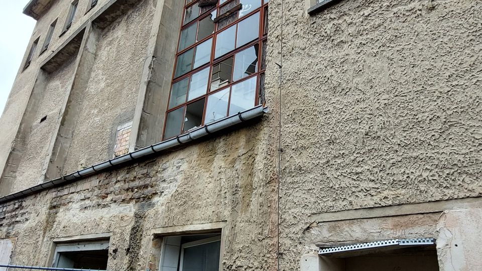 Ein mehrstöckiges Gebäude mit grober Putzfassade, kleinen rechteckigen Fenstern und einem großen Fenster mit roten Metallrahmen in der Mitte. Im Vordergrund ein einfacher Zaun, eine Metalltür und etwas Vegetation am unteren Rand der Fassade.