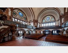 Innenraum einer historischen Kirche mit symmetrischen Rundbögen aus Backstein, Holzbänken, einer erhöhten Empore und bunten Rosettenfenstern. Die Decke ist gewölbt, und unter den Fenstern sind Statuen und dekorative Verzierungen sichtbar.