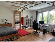 Ein Schlafzimmer mit Sport- und Arbeitsbereich. Links befindet sich ein Bett aus Holz mit Schubladen, daneben eine Sprossenwand mit Boxsack, Turnringen und Bodenmatte. Rechts steht ein Schreibtisch mit einem Computer, Bürostuhl und Regalen, und im Hintergrund sind große Fenster mit Blick ins Grüne.