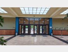 Geräumiger Eingangsbereich mit großen Glastüren, die nach draußen führen. Die Wände sind hell, teilweise blau abgesetzt, Holzelemente und Pflanzen säumen den Raum; die Decke ist mit einer gitterartigen Fensterkonstruktion gestaltet.