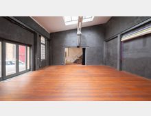 Großer, leerer Innenraum mit dunklen Wänden, einem Holzboden und einem Oberlicht. Links große Fenster- und Türfront, rechts mehrere aufgehängte Hintergrundrollen, hinten ein Durchgang zu einem Raum mit metallener Treppe.