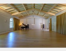 Ein heller, großzügiger Saal mit Holzfußboden und sichtbarer Deckenholzkonstruktion. An der Stirnwand stehen ein schwarzer Flügel und zwei Lautsprecher, die Wände sind mit Vorhängen und schlichten weißen Paneelen gestaltet.