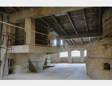 Innenraum einer alten Industriehalle mit Backsteinwänden, großen Fenstern und sichtbarer Holzbalkendecke. Im Vordergrund befinden sich ein runder gemauerter Ofen und eine Betonstruktur mit Treppenzugang, die in den Raum hineinragt.