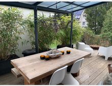 Terrasse mit Holzboden und einer Überdachung aus Glas und Metall, ausgestattet mit einem rustikalen Holztisch, weißen modernen Stühlen und dekorativen Elementen. Im Hintergrund befinden sich hohe Pflanzen in schwarzen Pflanzkübeln sowie ein Blick auf einen Garten mit Bäumen und einem Nachbarhaus.