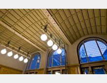 Innenansicht eines historischen Gebäudes mit einer hohen, gewölbten Decke, die mit filigranen Mustern und Zierleisten dekoriert ist. Große, bogenförmige Fenster mit Sprossengittern lassen blaues Tageslicht herein, während hängende Lampen mit kugelförmigen Leuchten den Raum erhellen.