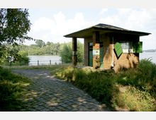 Backsteingebäude mit Flachdach und grünen Fensterläden und Türen, gelegen an einem gepflasterten Weg nahe einem Flussufer, umgeben von Bäumen und dichter Vegetation im Hintergrund.