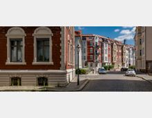 Straßenszene in einem Wohnviertel mit gepflasterter Straße, umgeben von mehrstöckigen Altbauten; links ein Gebäude mit rotem Klinker, rechts bunte, renovierte Fassaden im Stil der Gründerzeit. Im Vordergrund stehen Laternenpfähle, Autos und ein Fahrradfahrer sind zu sehen.