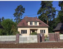 Zweistöckiges Einfamilienhaus mit beige verputzter Fassade und rotbraunem Ziegeldach, umgeben von einem Garten und einem weißen Lattenzaun mit Backsteinpfeilern. Im Hintergrund sind Bäume und ein weiteres Haus erkennbar.