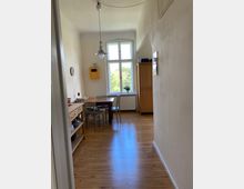 Schmaler Raum mit Holzboden und hohen Decken, eingerichtet als Essbereich mit einem Tisch, zwei Stühlen, einer Holzkommode und einem Schrank; im Hintergrund ein großes Fenster, das Tageslicht hereinlässt und den Blick auf grüne Bäume freigibt.