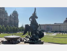 Eine Bronze-Statue zeigt einen Reiter auf einem Pferd im Kampf mit einem Löwen auf einem Platz mit Rasenflächen. Im Hintergrund sind der Berliner Dom, das rekonstruierte Berliner Stadtschloss und weitere historische Gebäude zu sehen, umgeben von Bäumen und Brunnen.