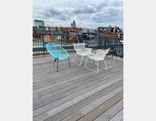 Dachterrasse mit Holzboden, darauf stehen zwei weiße Stühle und ein türkisfarbener Stuhl aus Metallgeflecht, im Hintergrund eine Stadtansicht mit flachen Ziegeldächern und einem Kirchturm unter einem bewölkten Himmel.