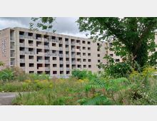 Verlassenes mehrstöckiges Gebäude mit offenen Fenstern und Graffiti an den Wänden, umgeben von wucherndem Grün und einzelnen Bäumen. Der Himmel ist bewölkt, und das Gelände wirkt ungenutzt.