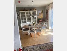 Esszimmer mit einem runden Holztisch, umgeben von weißen Stühlen mit gepolsterten Sitzflächen. Im Hintergrund steht ein weißer Vitrinenschrank vor einer grauen Wand, dekoriert mit Keramikgefäßen und Bildern. Rechts befindet sich eine große Fensterfront mit hellem Tageslicht.