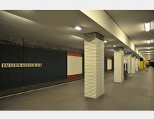 U-Bahnhof mit dem Schriftzug "Kaiserin-Augusta-Str." an der Wand, weißen Kachelsäulen, leeren Sitzbänken und beleuchtetem, schlichtem Design. Im Hintergrund sind Gleise und ein gelber Fahrkartenautomat erkennbar.