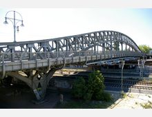 Metallbrücke mit geschwungenen Bögen und Geländern, die eine mehrspurige Bahntrasse überquert. Im Hintergrund sind Oberleitungen, Bäume und ein klarer Himmel sichtbar.