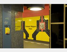 U-Bahn-Station mit gelb-schwarzen Wandfliesen, detaillierter Grafik und der Beschriftung „Richard-Wagner-Platz“. Ein Teil eines gepunkteten blauen Pfeilers und ein gelbes Fach mit Fahrplänen sind sichtbar.