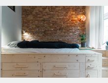 Holzbett mit eingebauten Schubladen vor einer Ziegelsteinwand, beleuchtet durch eine kleine Wandlampe und eine hängende Kugel-Pendelleuchte. Rechts befindet sich ein Fenster mit weißen Vorhängen, neben dem eine Pflanze auf einem runden Teppich steht.