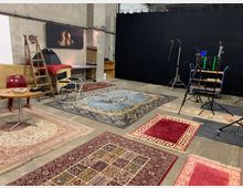 Eine Studio- oder Lagerhalle mit Betonwänden und einem schwarzen Vorhang im Hintergrund, ausgestattet mit mehreren orientalen Teppichen auf dem Boden. Im Raum befinden sich unter anderem eine Leiter, Stühle, ein Tisch, ein Monitor-Stativ und ein Regal mit Foto- oder Videozubehör; an der Wand hängt ein Bild eines Kaminfeuers.