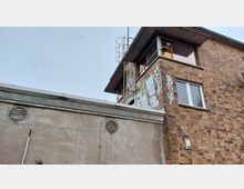 Backsteinfassade eines Gebäudes mit großen Fenstern und Graffiti, daneben eine Betonmauer mit Lüftungsgittern. Am oberen Rand des Gebäudes sind Metallleitern und Geländer angebracht.