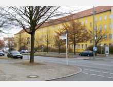 Gelbes Wohngebäude mit mehreren Stockwerken und roten Ziegeldächern, entlang einer Straße mit geparkten Autos und kahlen Bäumen. Im Vordergrund befindet sich ein Gehweg mit einem Straßenschild und einer Verkehrsinsel.