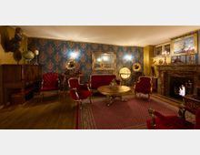 Ein elegantes Wohnzimmer im Vintage-Stil mit aufwendig gemusterten blauen Tapeten und klassischen Holzmöbeln. Zu den zentralen Elementen gehören eine rote Polster-Sitzgruppe, ein Kamin mit Marmorverkleidung, zwei Grammophone, ein Globus und ein ovaler Holztisch, umgeben von warmem Licht und Dekorationen im antiken Stil.