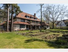 Großes Wohnhaus im Landhausstil mit rotem Ziegeldach und mehreren Gauben, umgeben von einem großen Garten mit Rasenflächen, alten Bäumen und Blumenbeeten. Im Vordergrund sind gestufte Gartenanlagen und tiefer liegende Beete erkennbar.
